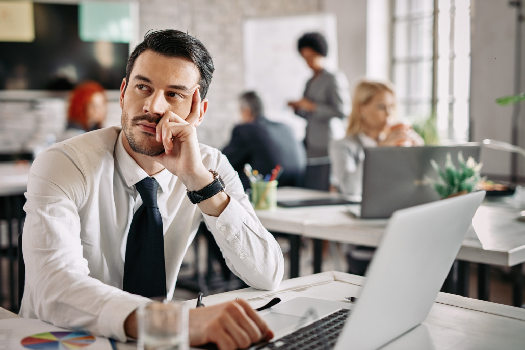 Business professional sitting at a desk in a modern office, looking thoughtfully to the side while working on a laptop, with coworkers collaborating in the background.